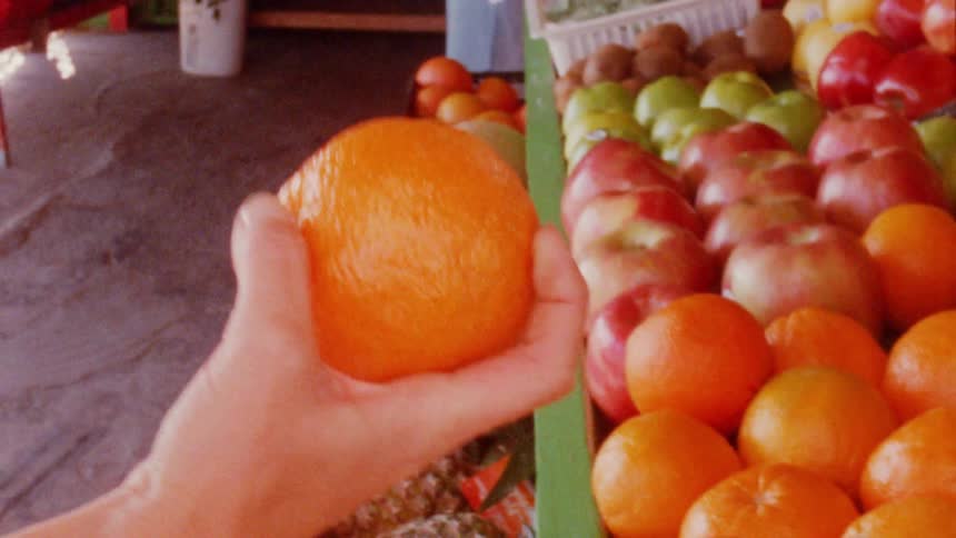 A woman picks up an orange and squeezes it at a beachside market