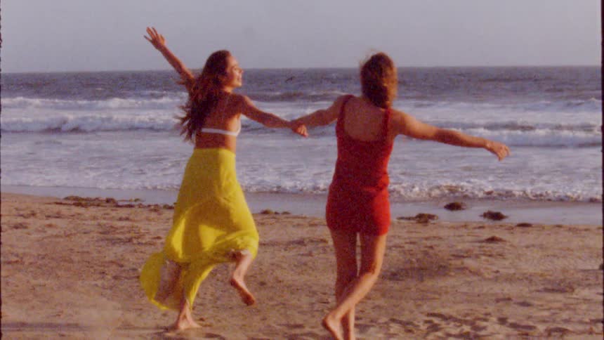 Two women hold hands and dance around on sandy beach as bird flies over ocean in background