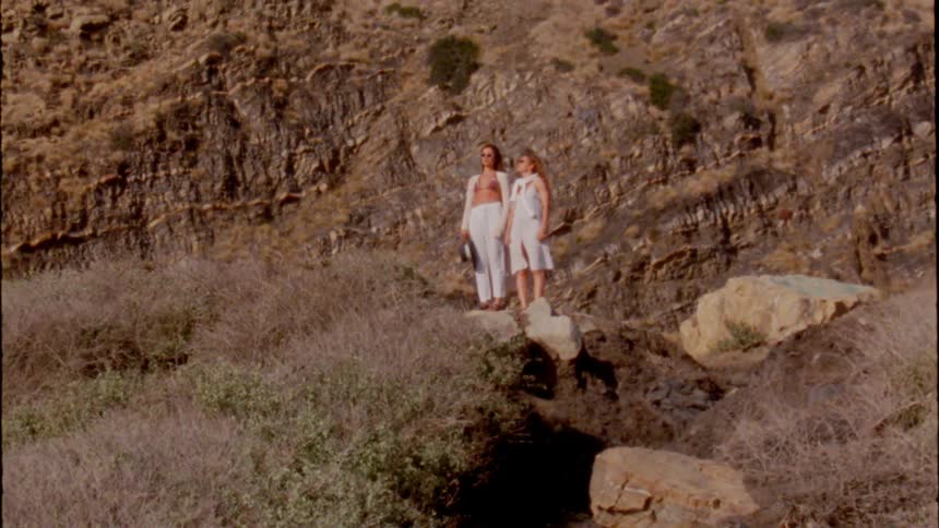 Two teenage girls wearing white clothes are standing on rocks on the side of a mountain and dancing