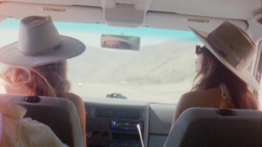 Two friends on vacation wearing sun hats driving a car while dancing