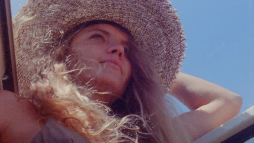 A woman wearing a bikini top and chain around her waist holds her sunhat on her head in the wind