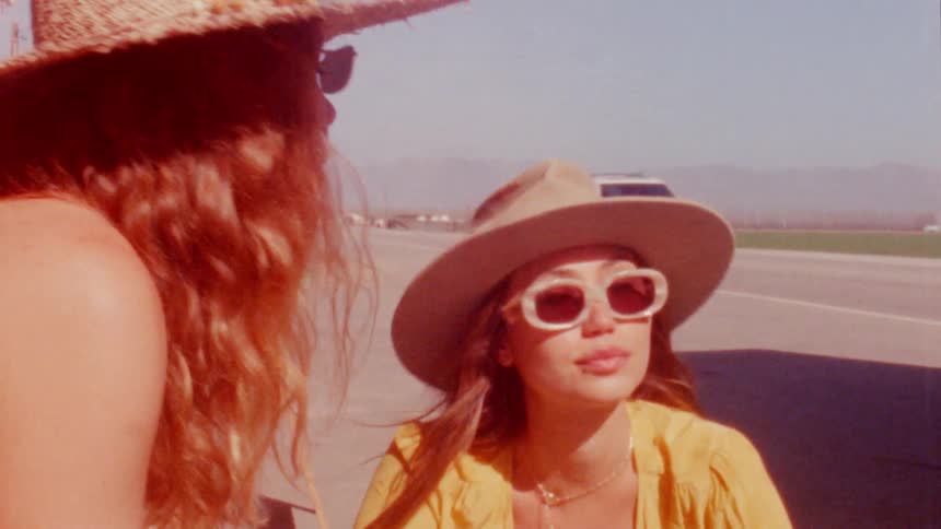 Woman waiting by side of road look at each other as traffic flows along highway with mountains on the horizon