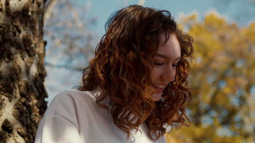 A teenage girl with curly red hair smiling while leaning next to a tree