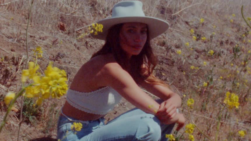 A girl wearing a white sun hat is sitting on a hill with yellow flowers