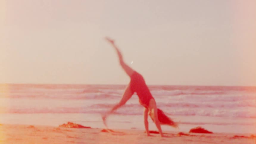 A woman does a cartwheel on a sandy beach with the ocean waves behind