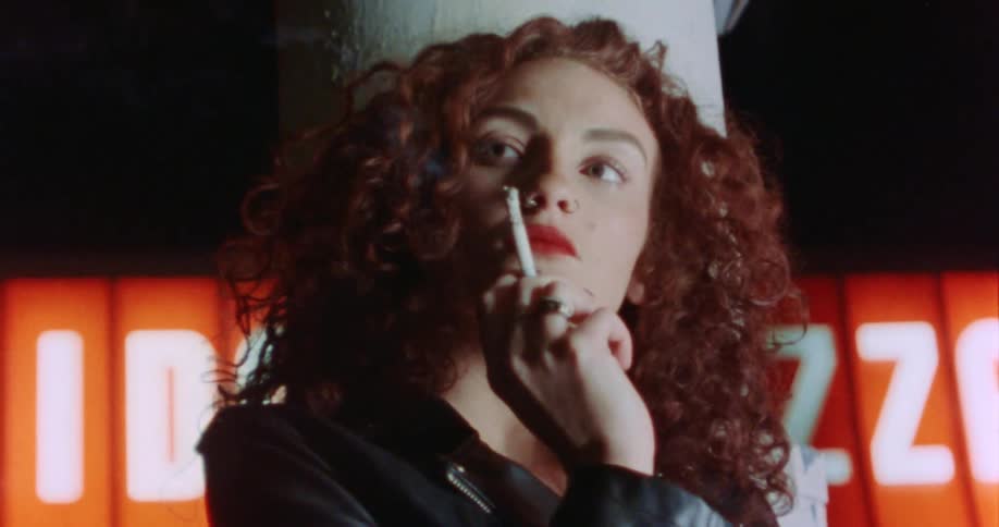 A woman poses holding a cigarette in front of a pizza shop at night