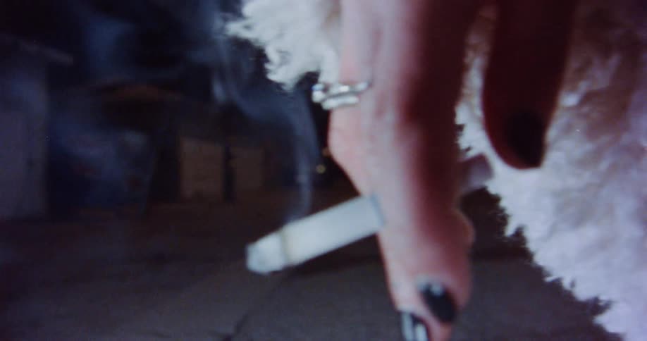 Smoke rises from a cigarette held by a woman wearing fluffy white clothing outside at night