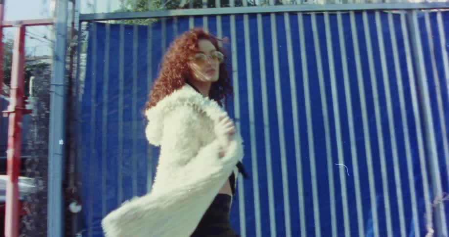 A woman wearing big glasses and red curly hair is wearing a fur coat and posing as she struts around