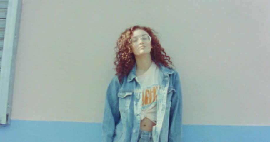 A woman wearing big glasses and red curly hair is standing against a wall modeling a denim outfit