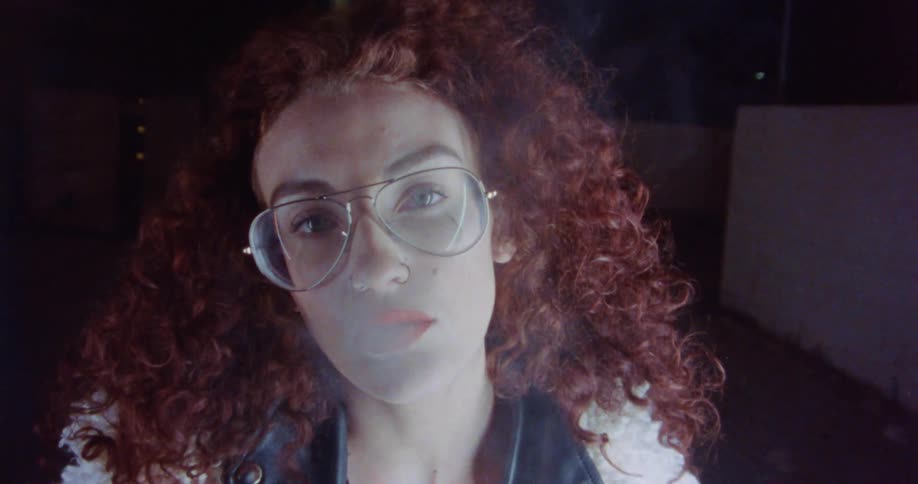 Redhead woman wearing glasses blows smoke from her mouth on the street at night
