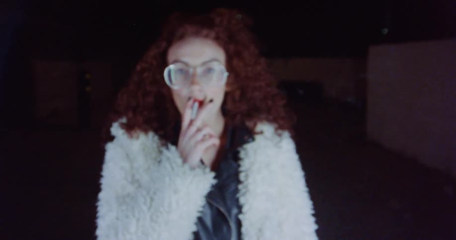 A woman wearing glasses is standing outside at night and smoking a cigarette