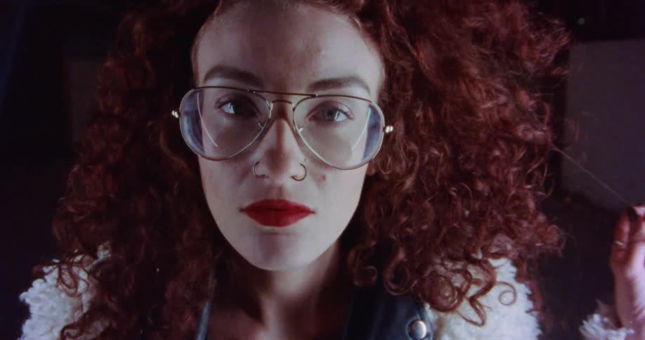 A woman wearing big glasses and red curly hair is looking forward and holing a strand of her hair in her hand