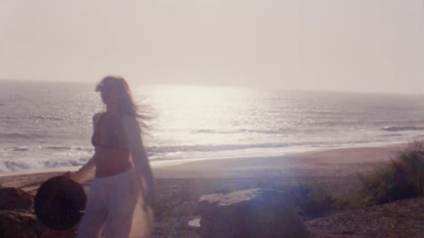 Two girls wearing white clothes are standing on a rock on a beach and then they jump down and walk away