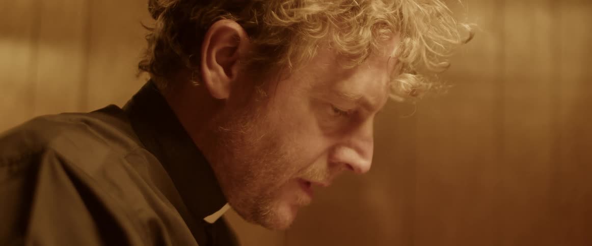 A priest leans forward as he stands with his head bowed