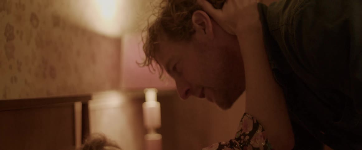 A woman is lying in bed and a man is standing over her as they hug and kiss
