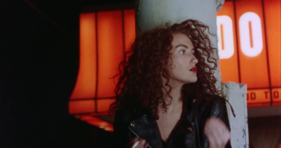 A woman standing against a pole in a parking lot ruffles her curly hair and smokes