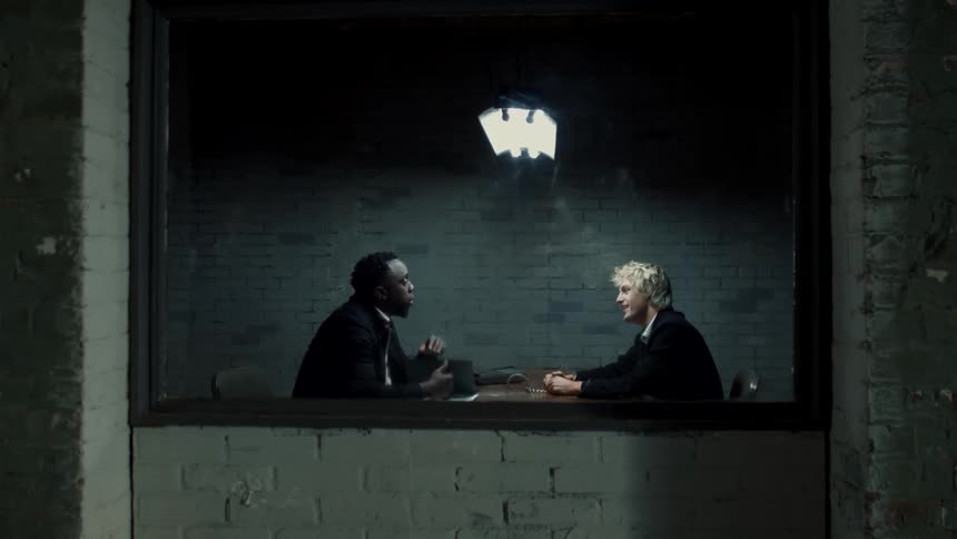 Two men sit at a desk inside a brick room and talk