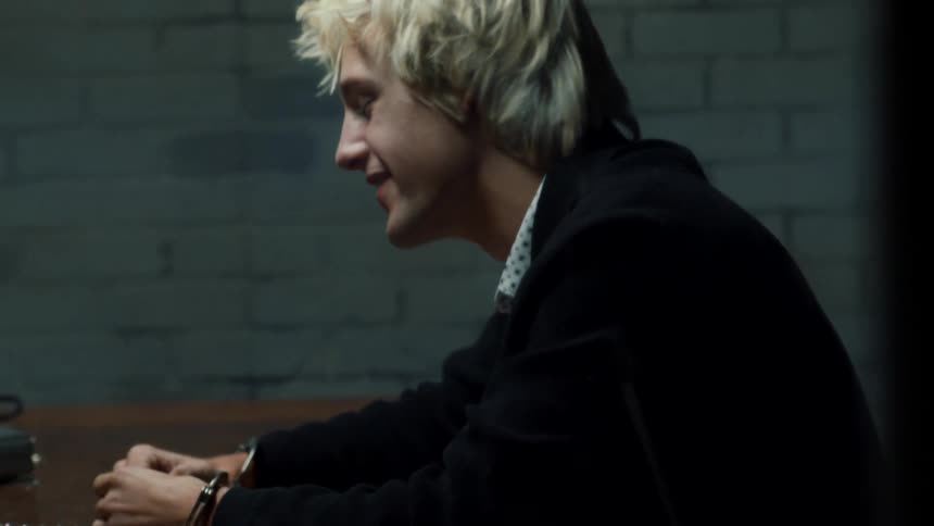 Blonde young man handcuffed to a table inside an interrogation room with white brick walls