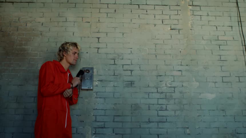 Young handcuffed man wearing prison orange overalls talking on a payphone against a white brick wall