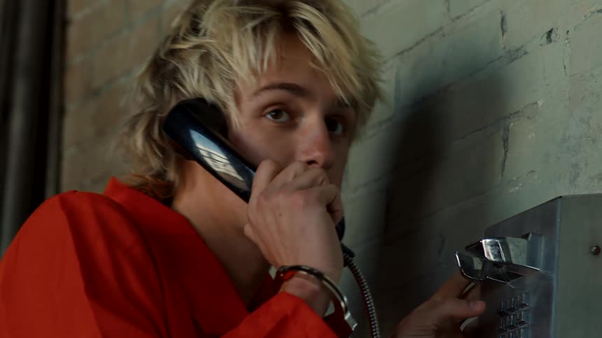 A pay phone is on a wall and a blond haired teenage boy picks up the receiver and makes a call