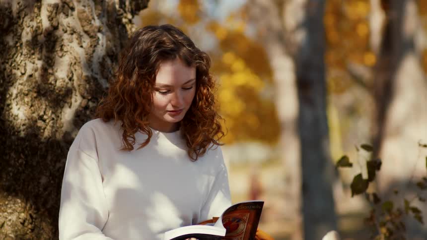 A woman is sitting up against a tree readinga book