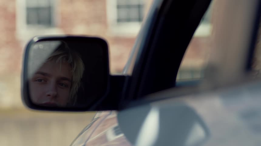 A mans face is reflected in a cars side mirror as it sits parked near a building