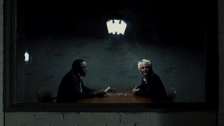 A man sitting at a table excited by the two way mirror as a detective tries to get his attention