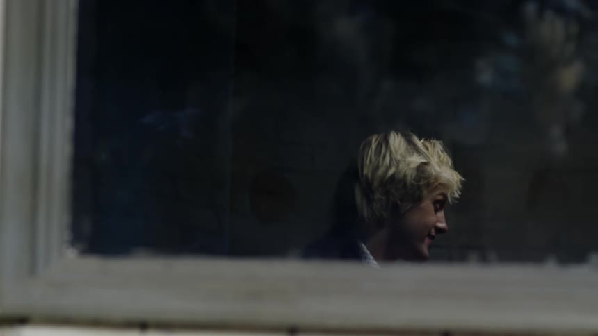 Young blonde man talking to another person stands up from the table seen through a window