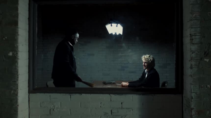 Handcuffed young blonde man looks up to greet a detective inside a interrogation room