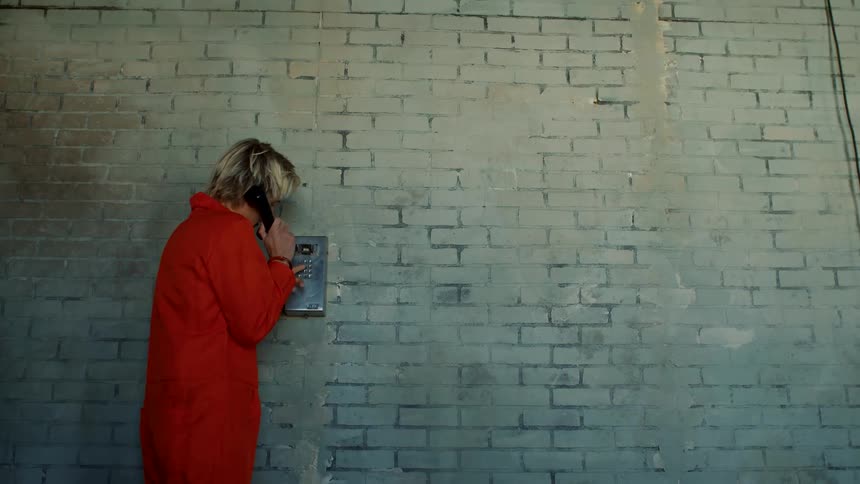 A handcuffed man in an orange jumpsuit walks to a phone on a wall and makes a call