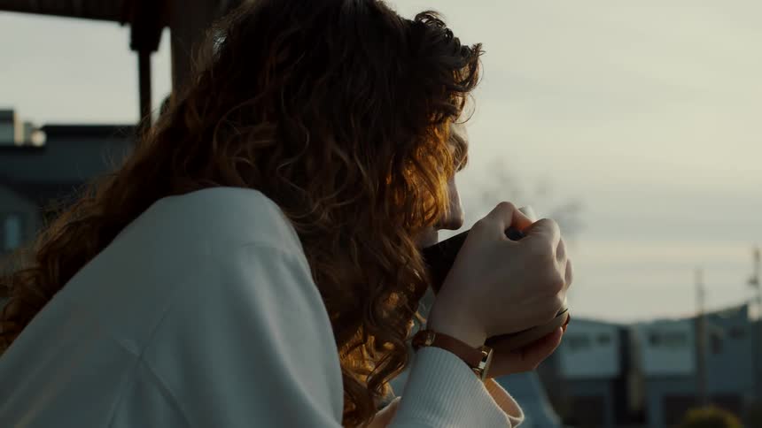 Young redhead woman drinking a hot beverage reclined on a balcony at sunset