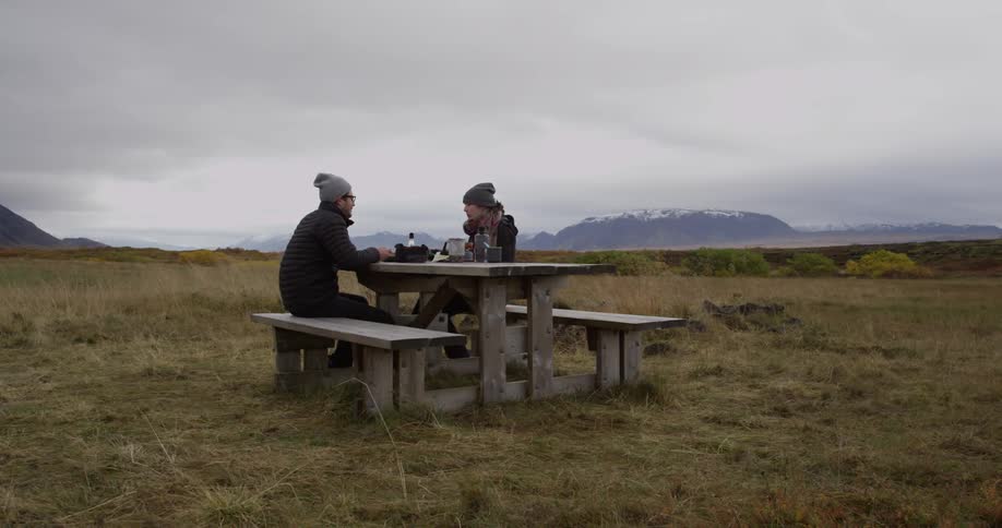 Two people sit down at a bench eating in the wilderness