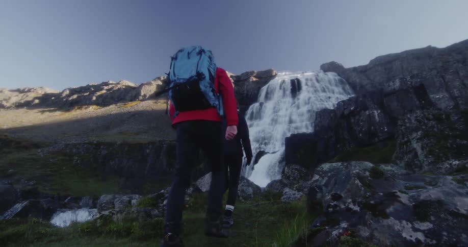 Two people hike uphill towards a large waterfall