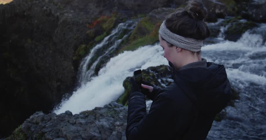 A woman is standing on the edge of a waterfall taking a photo with her phone