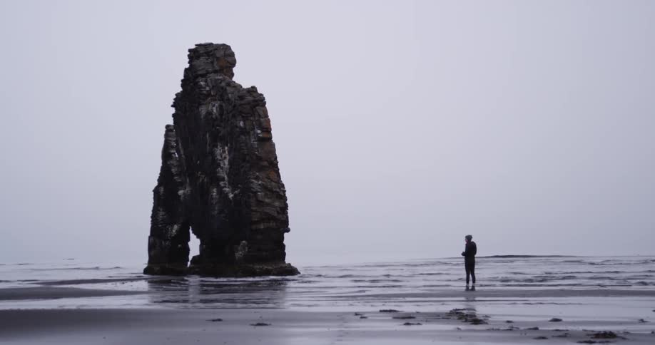 A person is standing before a large rock formation in the beach
