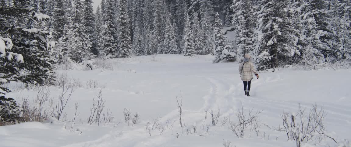Woman with beige beanie coat black pants and boots walks in snowy forest