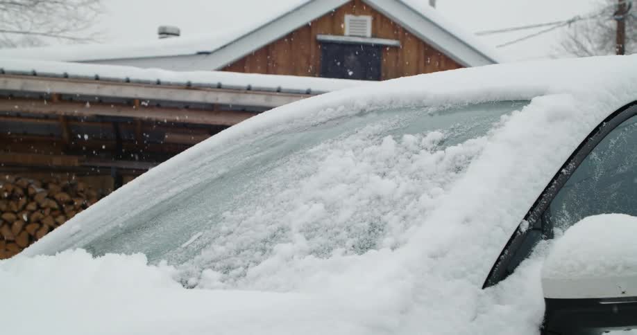 Wipers removing the accumulated snow on the windshield of a car parked in front of a cabin