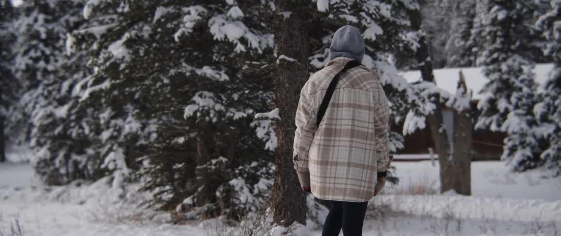 Woman with grey hoodie brown coat black pants walks in the snowy field next to country house
