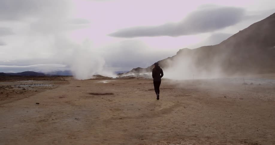 A woman walks along a deserted area with smoke rising in the distance