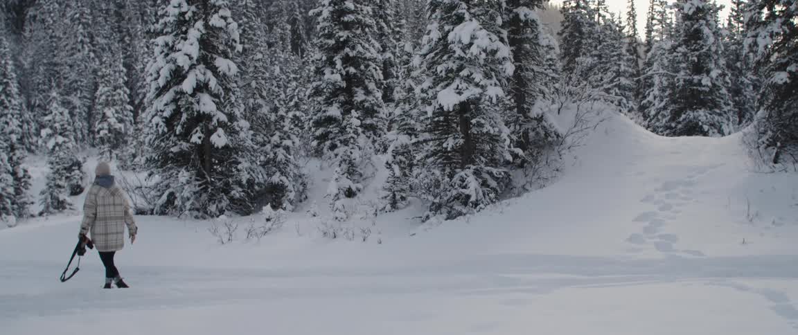 Woman with white beanie coat black pants and boots walks in snowy forest
