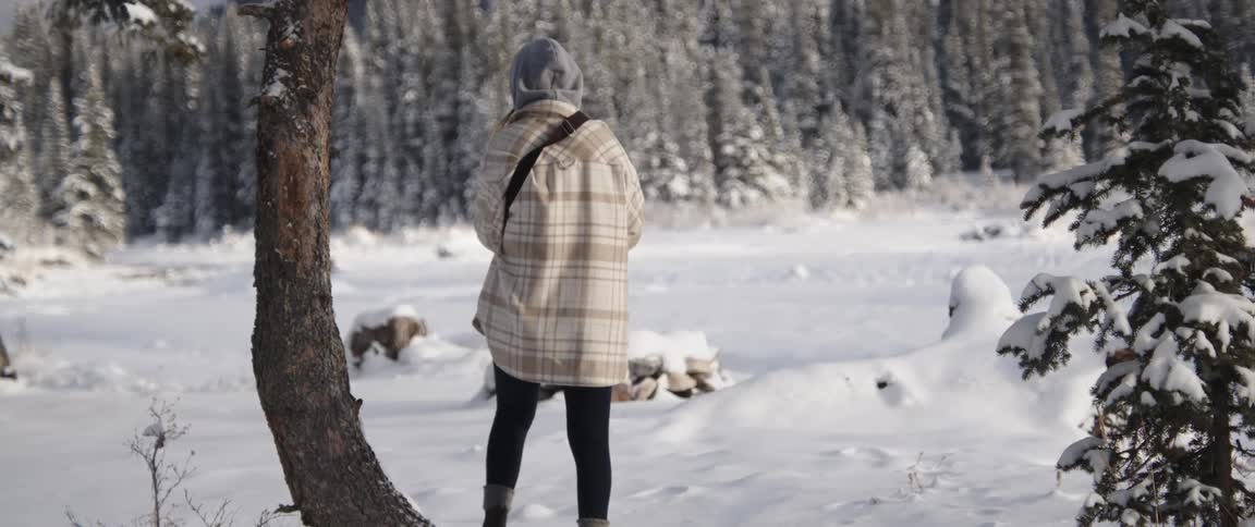 Woman standing in snow covered clearing in a conifer forest puts on mittens then turns to walk across snow