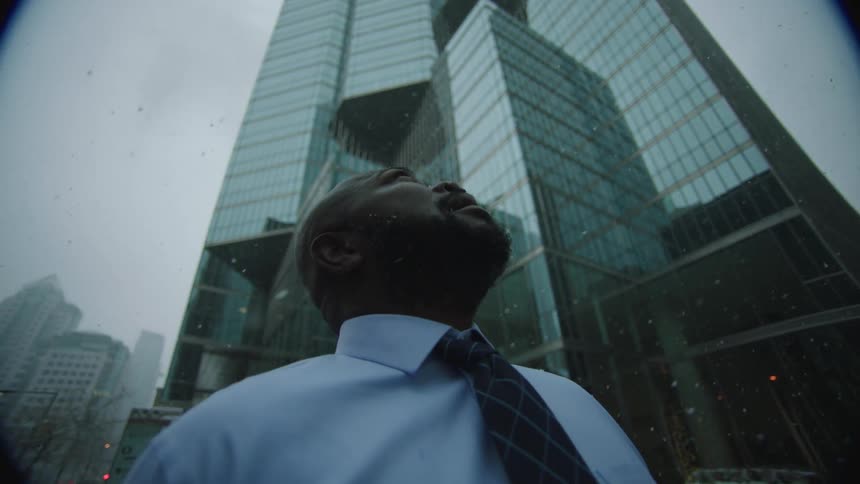 A man is standing on a city sidewalk and looking up as snow falls onto his face