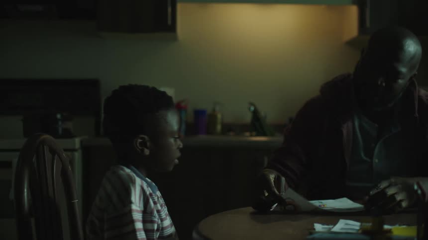 A man sits at a table and his son runs up to him and gives him a drawing and the man looks at it and nods and talks to his son