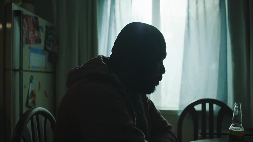 Sad bald man with beard and hoodie sits at table holding beer bottle in kitchen at home