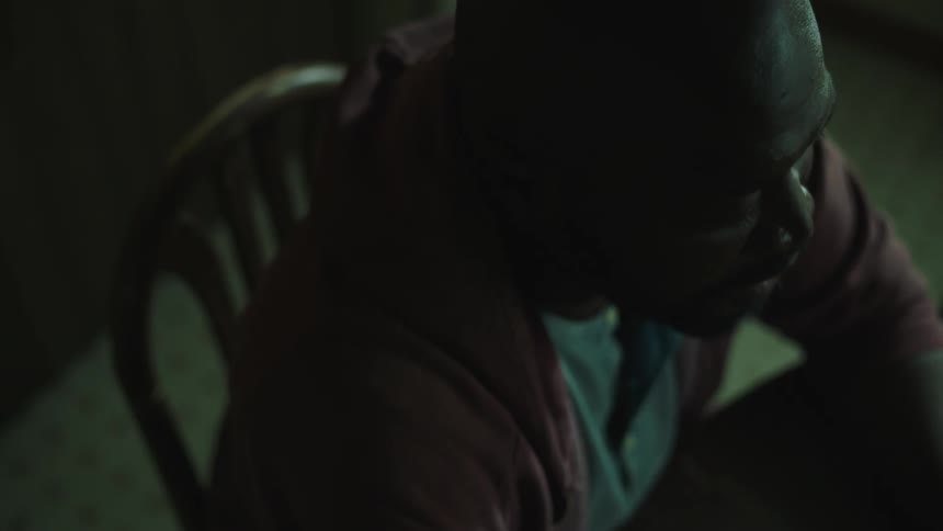 A black man is sitting in a chair in a room with dim light speaking to an unseen person