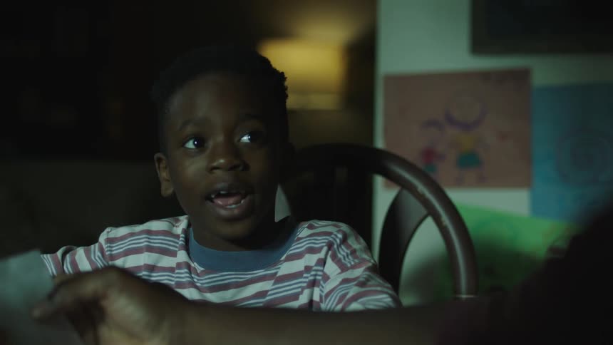 A young black boy sits down in a chair and shows someone sitting at the table a paper