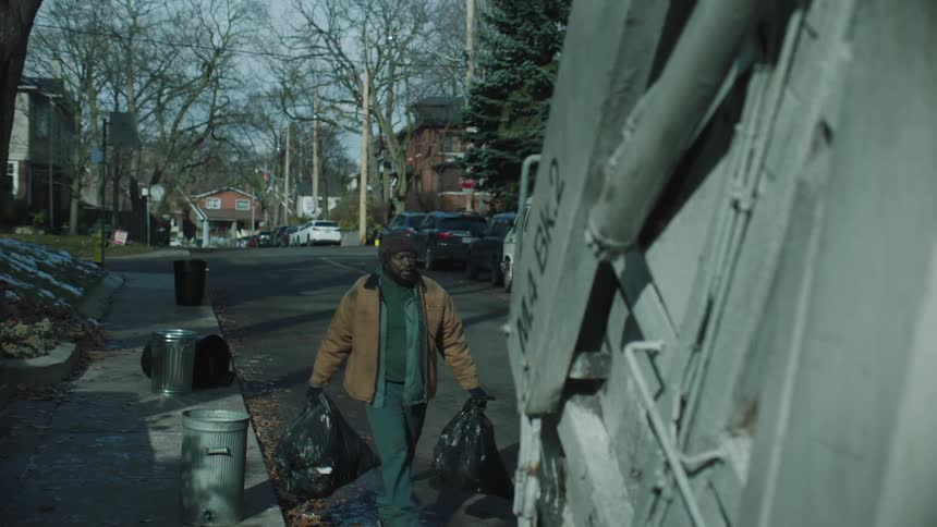 A male employee gets off of a garbage truck and picks up the garbage on the road to put in the truck
