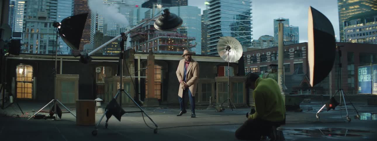 A black man is on a stage modeling a suit and there are tall city buildings behind the stage