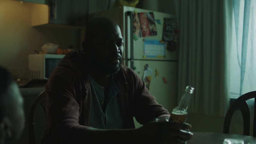 A man sits in a kitchen holding a beer bottle and talking to someone else