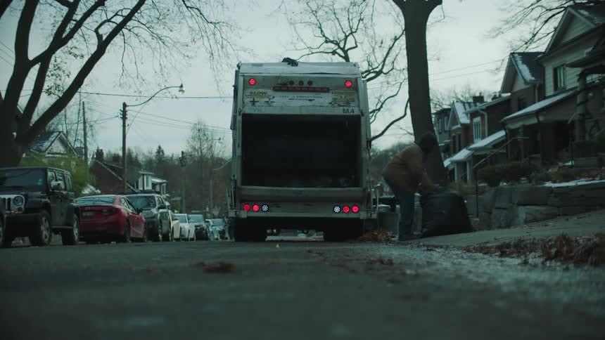 A garbage collector grabs trash bags and tosses them in the back of the truck before getting back on